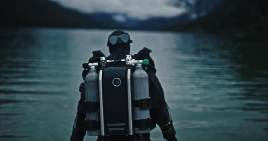 A man wearing diving gear drags two black bags into a lake at evening time
