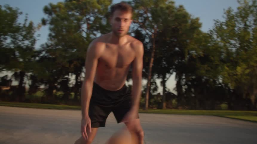 Topless basketball player wearing black shorts plays basketball in basketball court at sunset