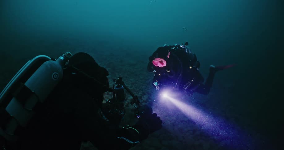 Two men wearing scuba gear at the bottom of the ocean are setting up camera equipment to film something on the ocean floor