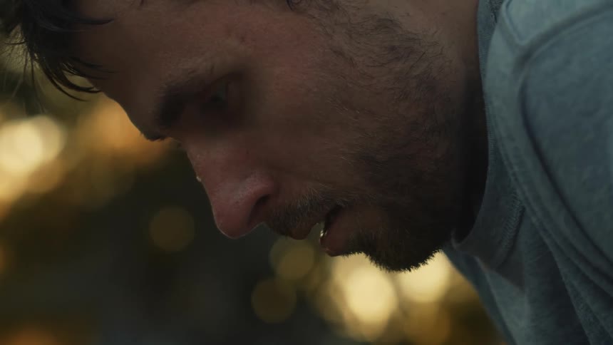 Man with beard and gray sweatshirt sweats and breathes with hands on his knees