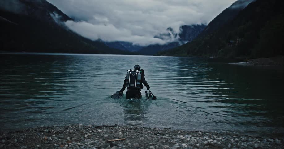 A man in full diving gear walks into the water of a mountain lake
