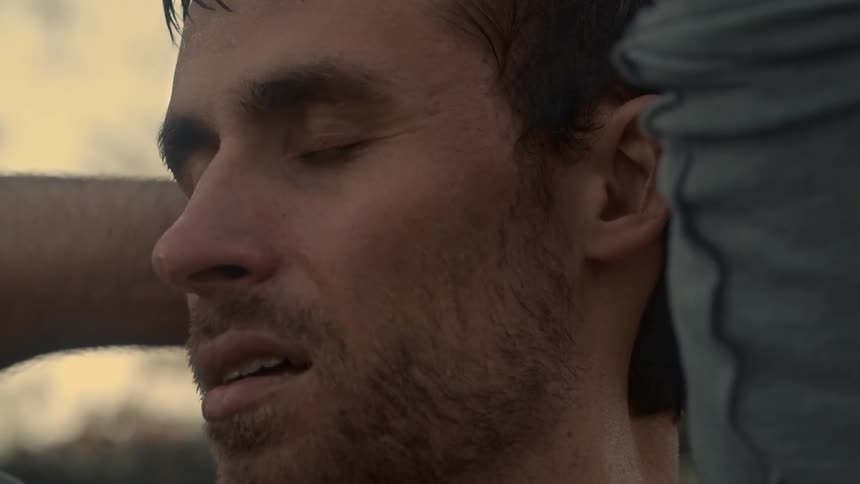 A man takes a deep breath while standing outside with his hands behind his head