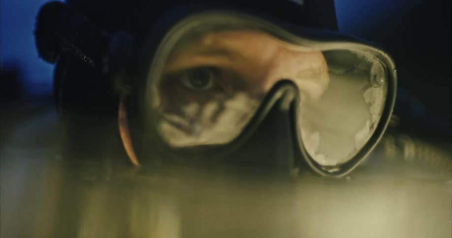 A close up of a mans face in scuba gear wearing a scuba mask shows him briefly going underwater and coming back up staring in one spot and looking depressed the whole time