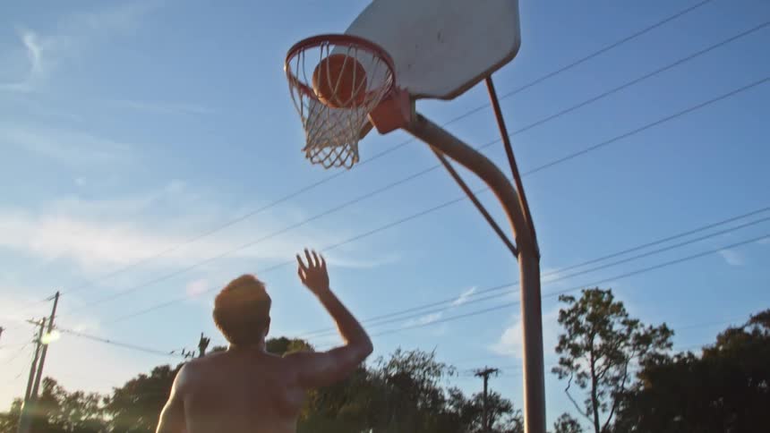 Topless man with black shorts plays basketball at sunset in basketball court