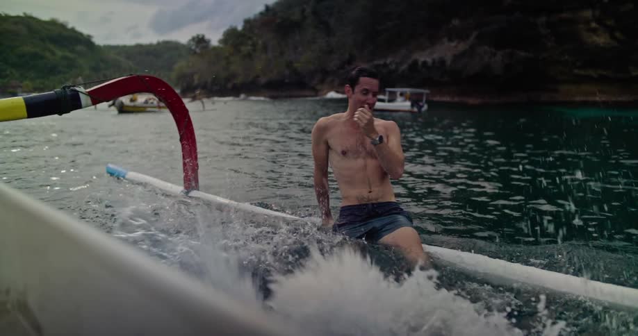 Young muscular shirtless man sitting on the pontoon of a boat close to a coastal beach watchs a friend jump on the water