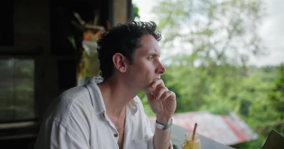 A man is sitting at a patio counter typing on his laptop computer and he stops to think