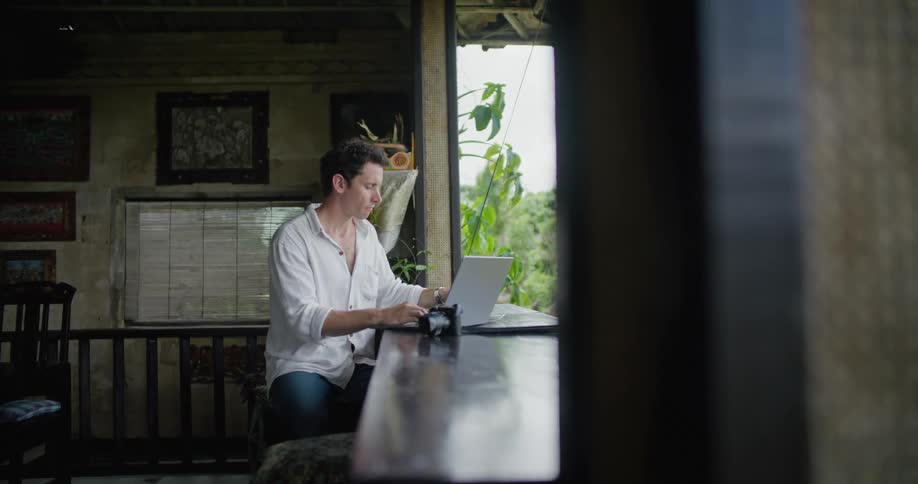 A man is sitting down at a counter with a laptop computer as he drinks his coffee and types on the keyboard and looks outside