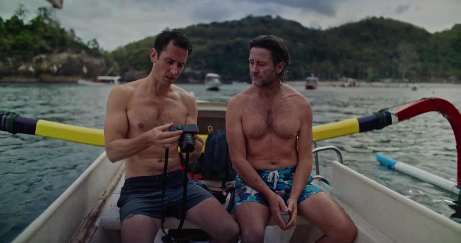 Couple of shirtless friends sitting on a boat near the coast looking at pictures on a camera