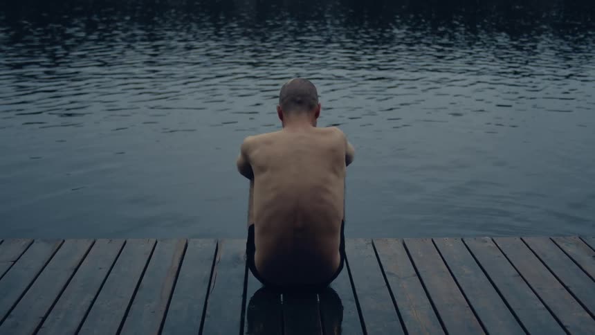 A man is sitting on the pier with his arms wrapped around his knees as he watches the water