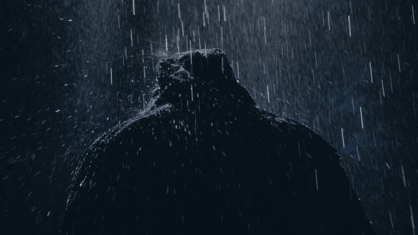 Rain falls and splashes on the black hooded jacket of a person with their back turned as it rains at night