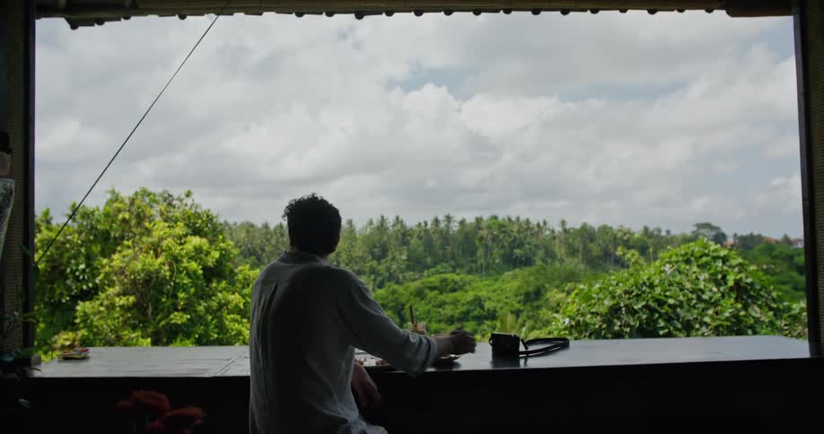 Photographer sitting on the balcony of a restaurant on the tropics drinks coffee while looking at the landscape