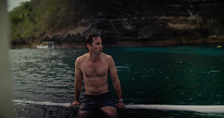 Young muscular shirtless man sitting on the pontoon of a boat close to a coastal beach