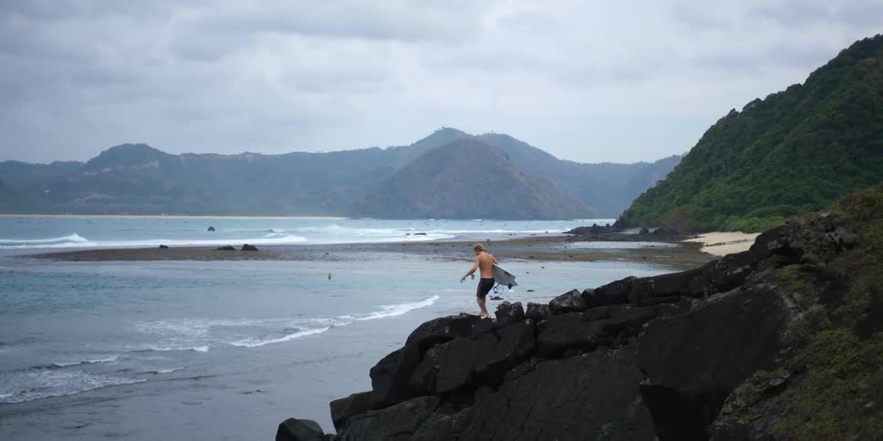 A man holding a white surf board running down a slope of rocks towards the ocean during the day