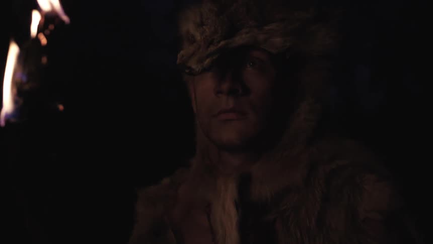 A person wearing a wolf carcass as a hat stands holding a flaming torch at night