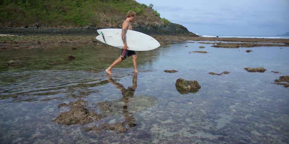 A man carrying a surfboard is walking into the clear water along the beach under a cloudy sky