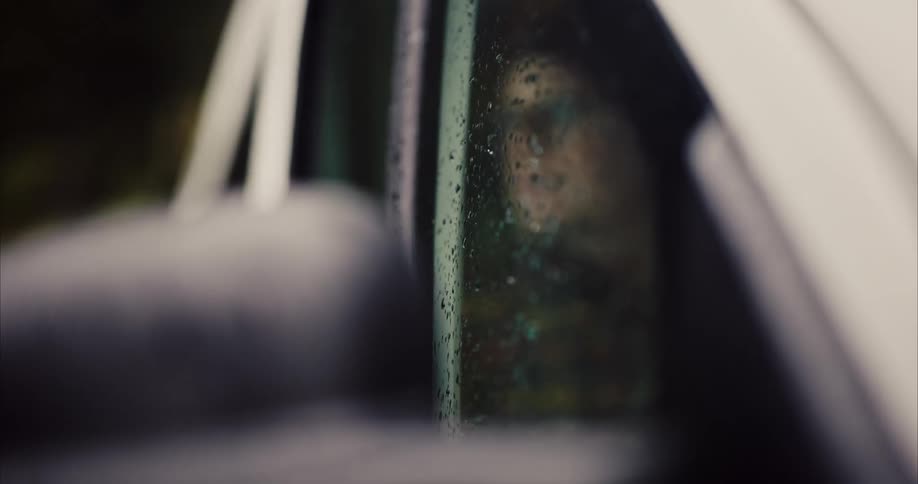 Face of a man wearing a beanie and glasses chewing bubblegum behind a wet glass