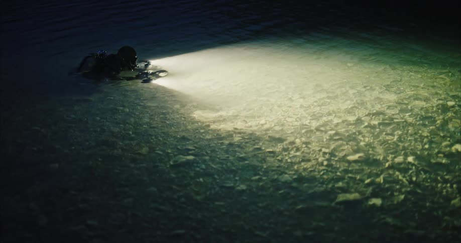 Scuba diver swims partly above water using light to scan rocky bottom under shallow water