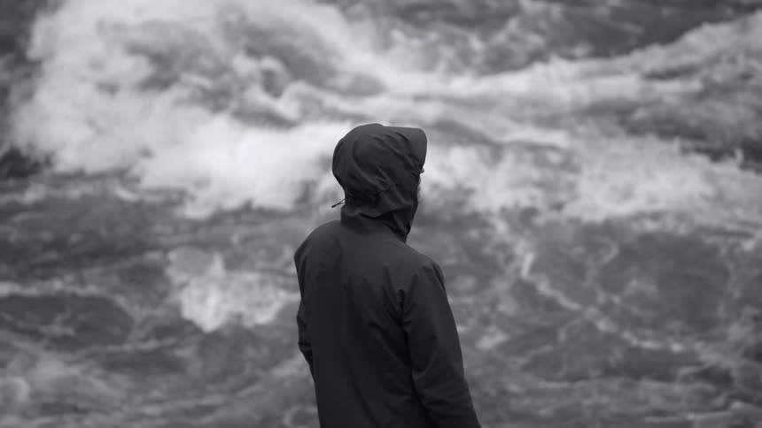 A person wearing a black hooded jacket is standing along the coast watching the rough water waves