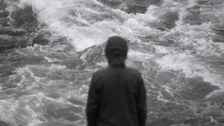 A blurry man in a black raincoat stands in the foreground in front of fast flowing river