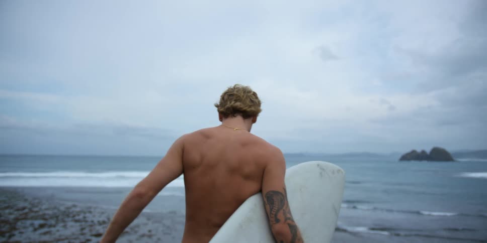 A man is walking on the beach holding a surfboard with his back to the camera