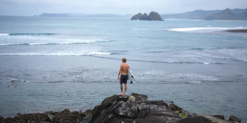 A man holding a surfboard hops onto the top of a pile of rocks on the coast
