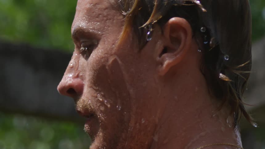 A man splashes water on his face from a bucket of water while standing outdoors
