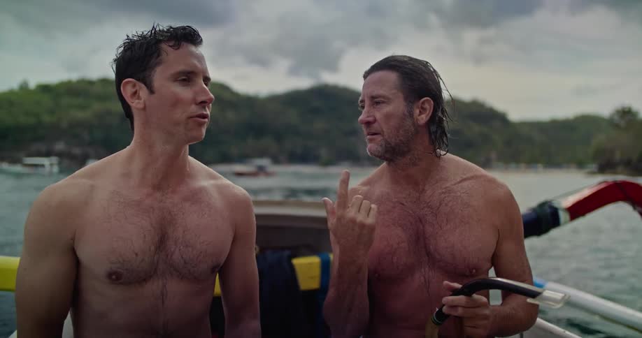 Shirtless friends sitting on a boat prepare to wear diving equipment near the coast