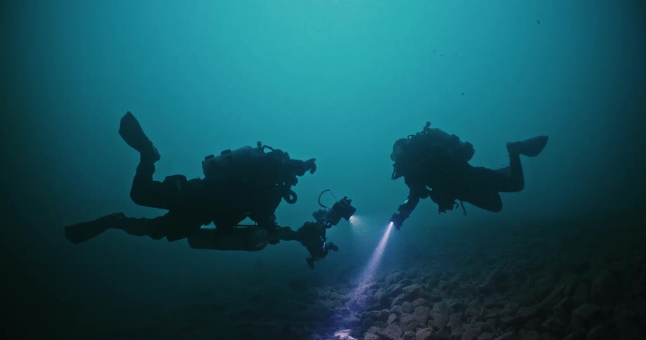 Two scuba divers silhouetted against murky water shine light and point camera at rocks on seafloor