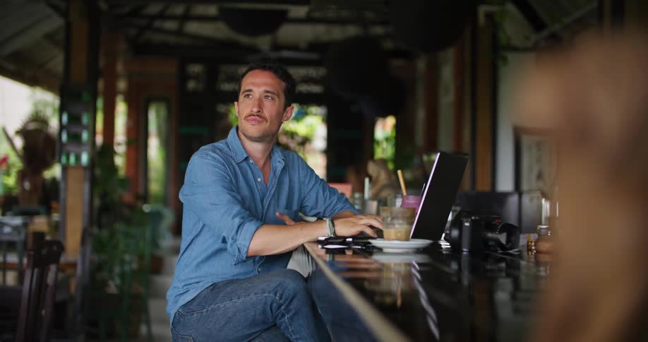 A man sits at a bar with a drink and types on a laptop