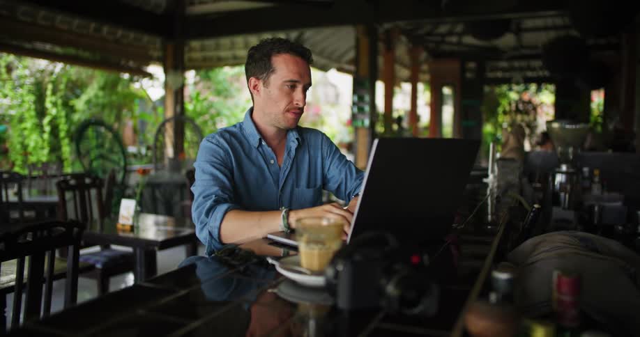 A man is sitting at a table having a drink as he types on his laptop computer