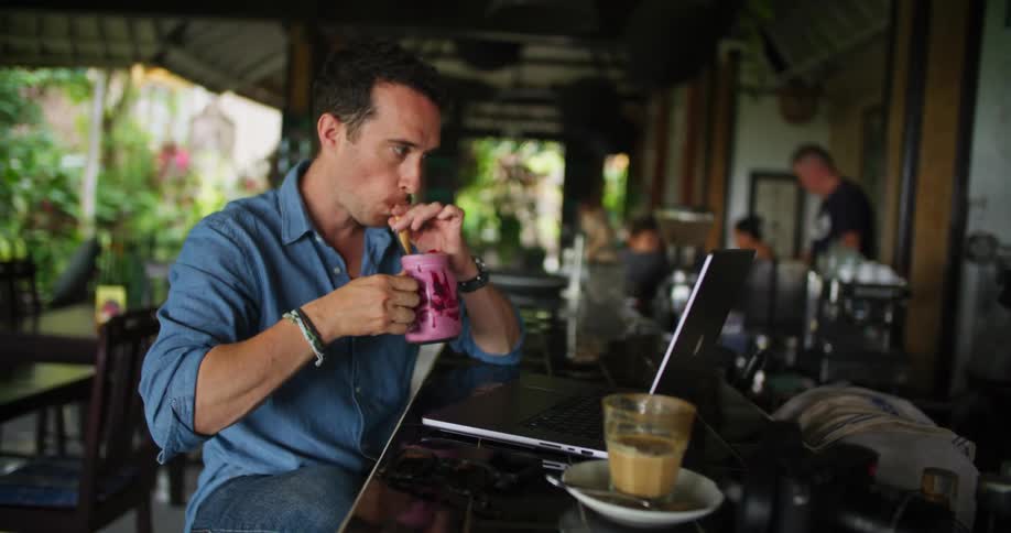 A man sits at a bar and takes a drink and types on his laptop