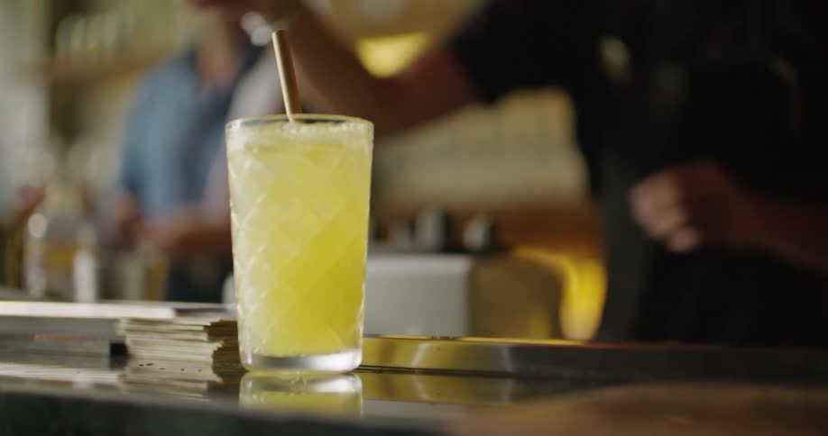 A drink is sitting on the bar and the bartender places a straw and small fruit in the glass