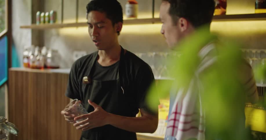 Two bartenders are talking behind the bar as one pats the other on the back as he reaches for a glass