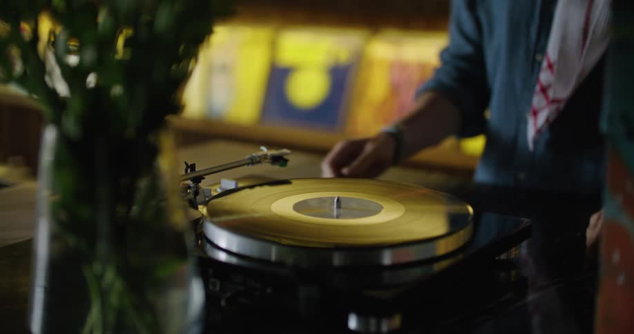 Man working in pub with several albums on the shelves under the stacked glasses places a record on then starts the record player