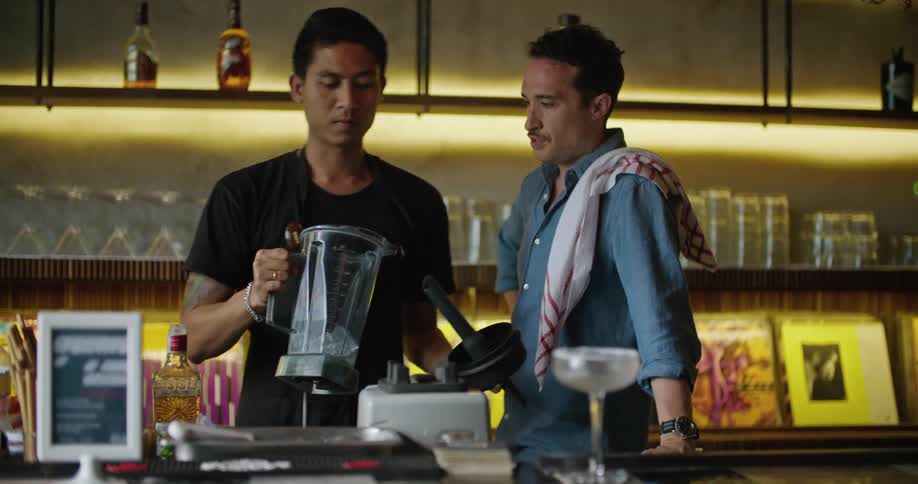 Bartender making a drink on the blender at the bar counter while chatting with a friend next to him