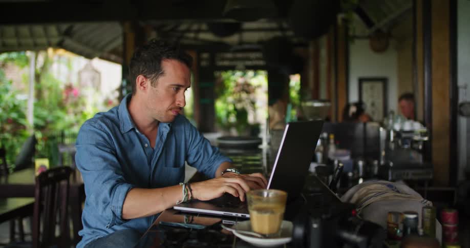A man sits at a bar and takes a drink and types on his laptop
