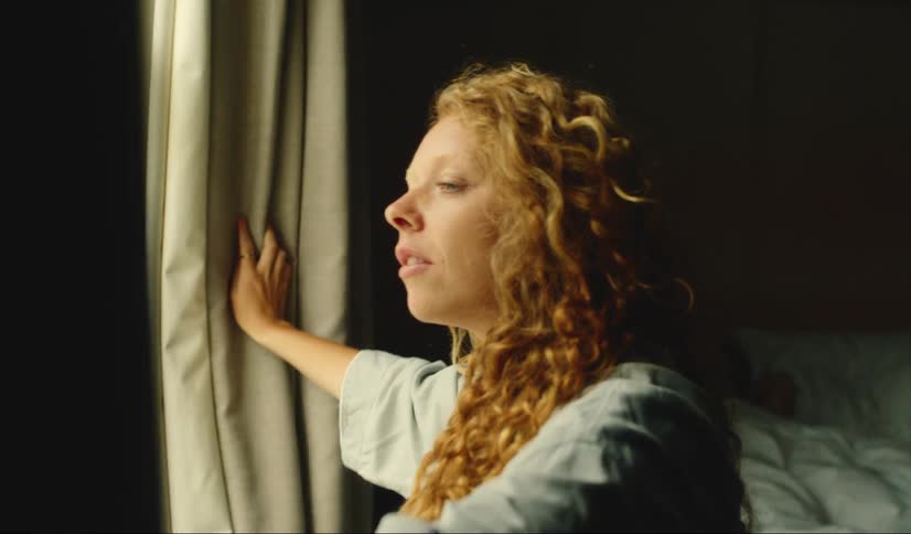 A woman opens up her drapes and looks outside and smiles