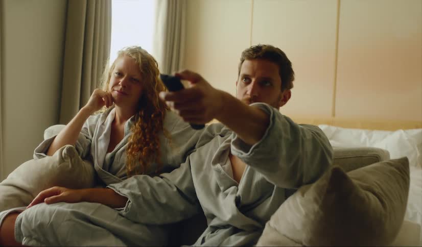 A man and woman lounging on a sofa in bathrobes and watching tv in hotel room