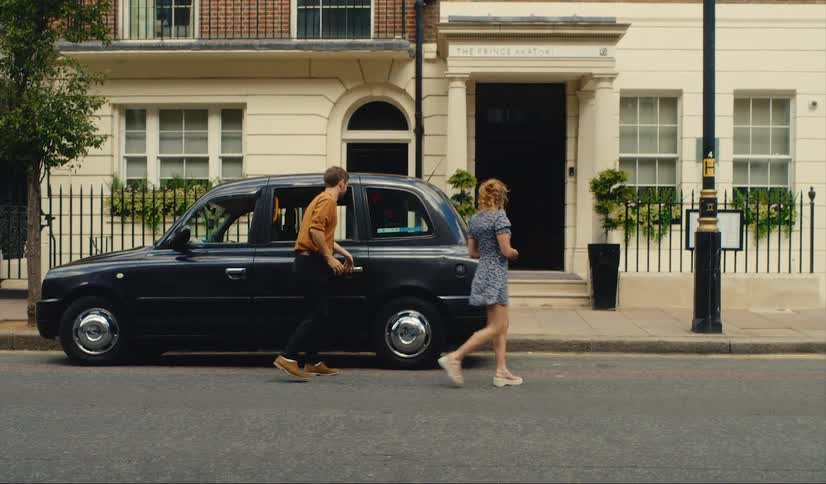 A man and a woman get out of a parked london taxi and he closes the door and puts his arm around her as they walk to a building