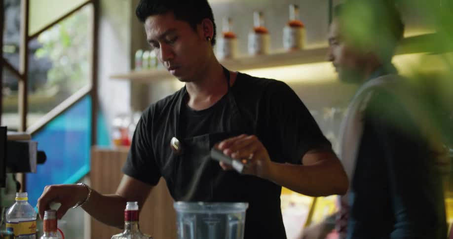 A bartender pours alcohol in to a blender as he talks with his coworker who then goes and wipes an espresso machine