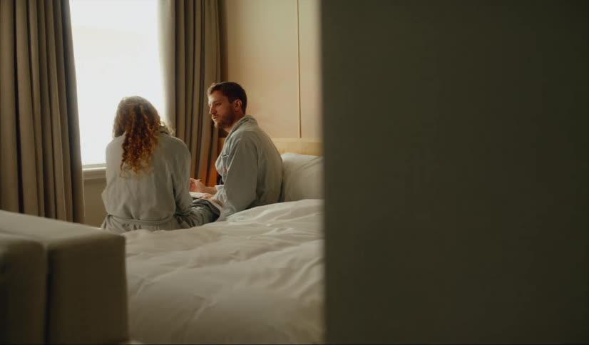 A man and woman sitting on a bed wearing matching bathrobes talking and sipping coffee