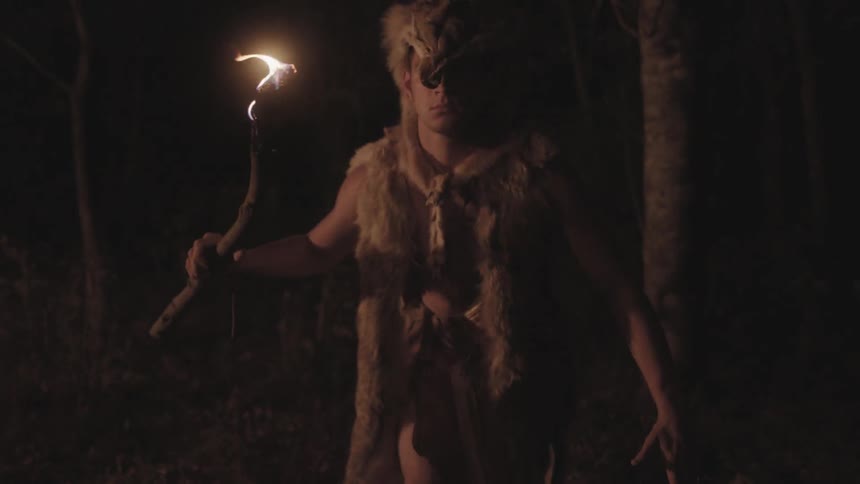 A man wearing clothing made of hide creeps through a forest at night