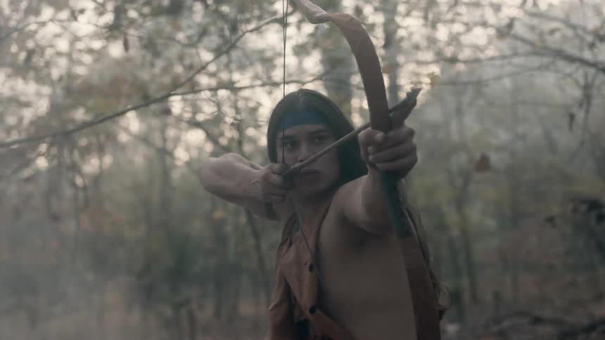 A native american standing in the woods with his bow and arrow drawn ready to shoot