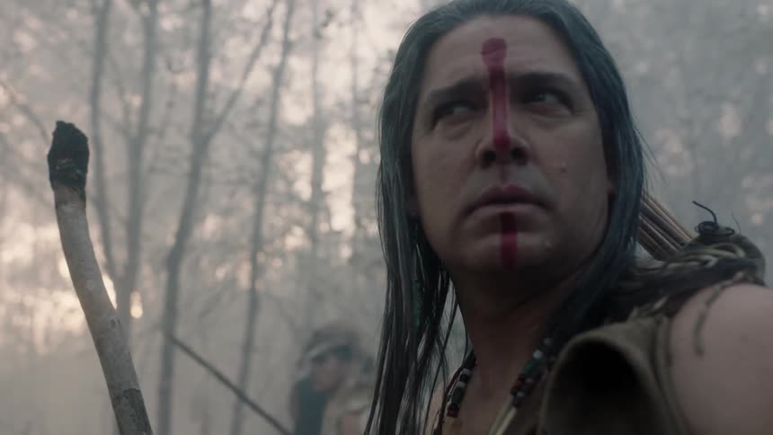 A native american carrying a bow stands looking around suspiciously in a misty forest
