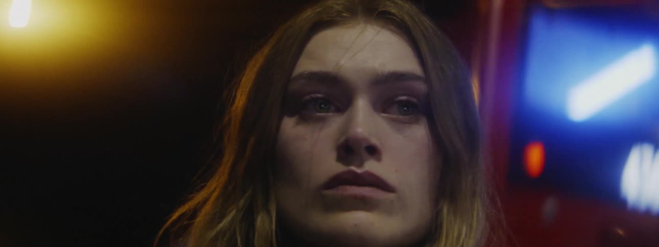 A sad looking woman stands outside at night as a bus drives behind her
