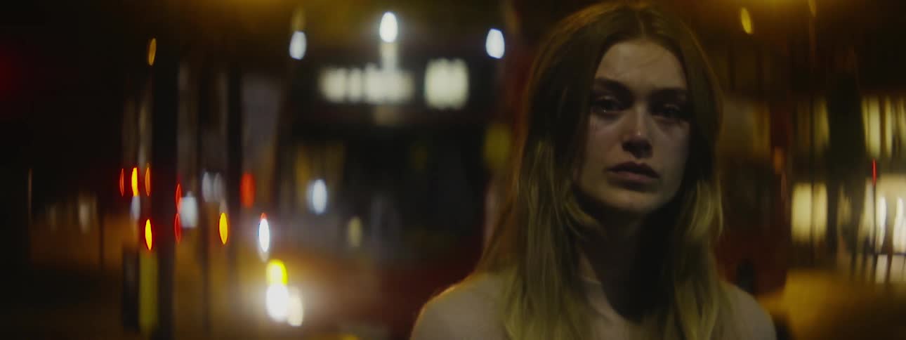 A nervous looking woman stands by a street at night as a double decker bus drives past