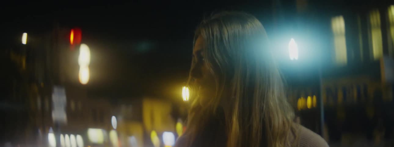 A woman is standing outside on a downtown street at night as the lights on buildings glow in the darkness