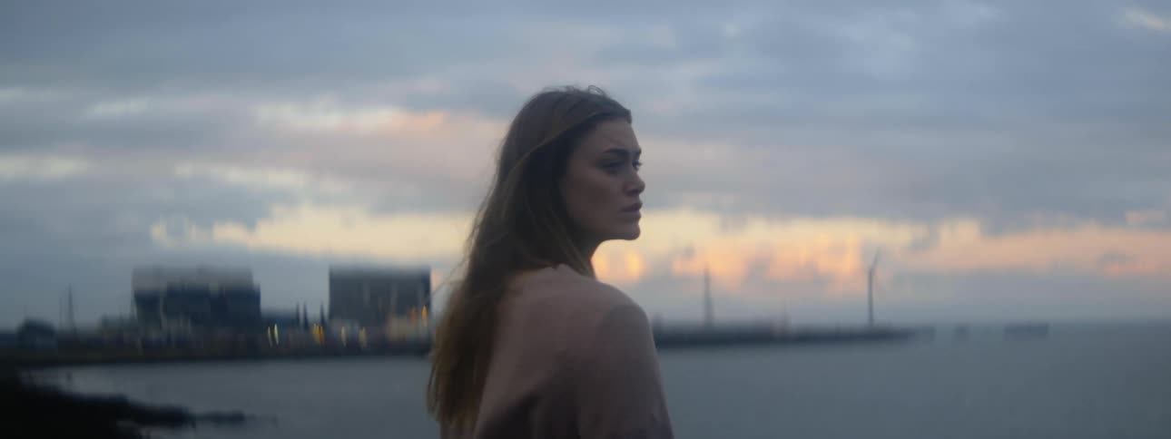 A woman is standing along the coast looking at the water and there are buildings near the pier