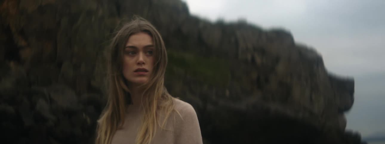 A woman is standing along the coast near rock formations with a concerned look on her face