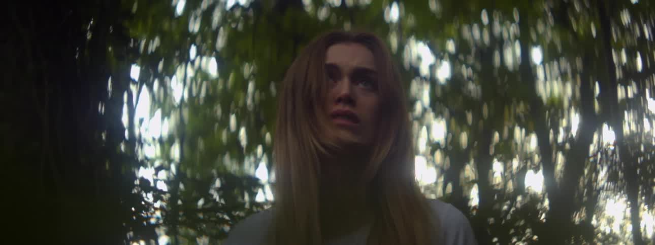 Woman with long hair stands under trees staring with a slight frown